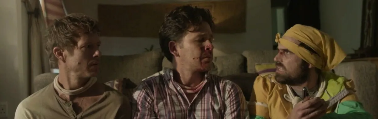 Three men sit side by side on a couch, one with a bloodied nose